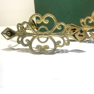 Puede incluir: Adorno decorativo de metal color lat&oacute;n ornamentado, con un extremo en forma de diamante y detalles de ojo de cerradura. El dise&ntilde;o presenta patrones arremolinados y una est&eacute;tica vintage. Una caja verde est&aacute; en el fondo.