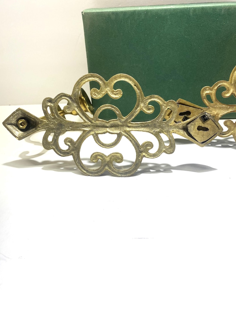 Puede incluir: Adorno decorativo de metal color lat&oacute;n ornamentado, con un extremo en forma de diamante y detalles de ojo de cerradura. El dise&ntilde;o presenta patrones arremolinados y una est&eacute;tica vintage. Una caja verde est&aacute; en el fondo.