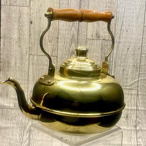 May include: A vintage brass kettle with a wooden handle. The kettle has a rounded body, a curved spout, and a lid with a small knob. The handle is made of light brown wood. The kettle is sitting on a clear surface.
