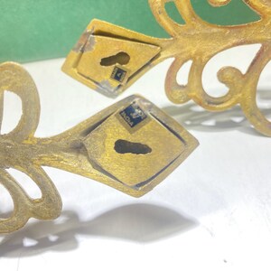 Puede incluir: Primer plano de dos soportes de pared ornamentados de metal dorado. Cada soporte presenta un dise&ntilde;o decorativo en espiral y una placa de montaje en forma de diamante con un ojo de cerradura. Los soportes est&aacute;n marcados "Made in India".