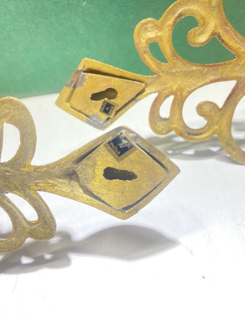 Puede incluir: Primer plano de dos soportes de pared ornamentados de metal dorado. Cada soporte presenta un dise&ntilde;o decorativo en espiral y una placa de montaje en forma de diamante con un ojo de cerradura. Los soportes est&aacute;n marcados "Made in India".
