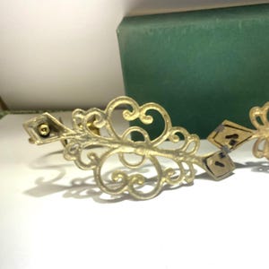 Puede incluir: Pieza decorativa de metal dorado ornamentada con un dise&ntilde;o en espiral. La pieza tiene detalles en forma de diamante y un peque&ntilde;o elemento circular. Un objeto rectangular verde se encuentra en el fondo.