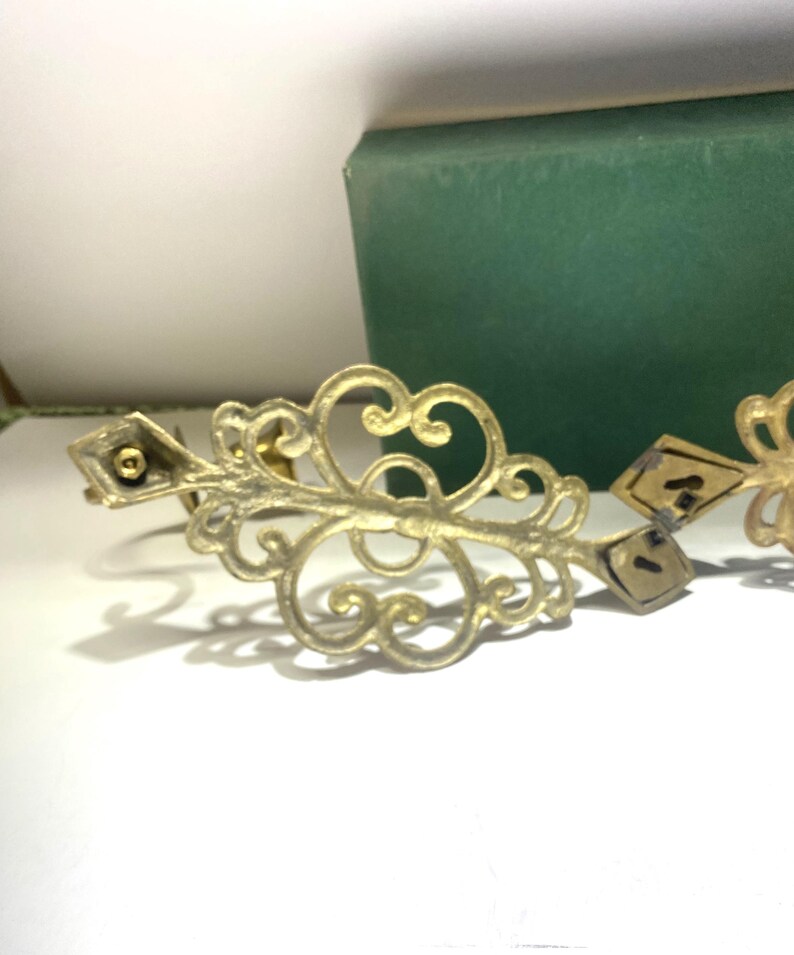 Puede incluir: Pieza decorativa de metal dorado ornamentada con un dise&ntilde;o en espiral. La pieza tiene detalles en forma de diamante y un peque&ntilde;o elemento circular. Un objeto rectangular verde se encuentra en el fondo.