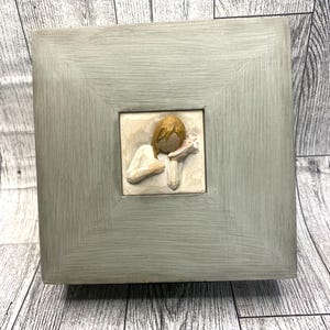 May include: A square, gray-painted wooden box with a framed, raised-relief sculpture of a person holding a flower. The sculpture is in a square, off-white tile. The box is sitting on a weathered, gray wooden surface.