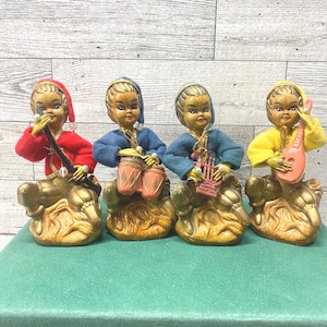 May include: Four vintage figurines of musicians. Each figure is dressed in a different colored outfit: red, blue, blue-green, and yellow. They are seated on gold-colored bases. Each figure is playing a different instrument.