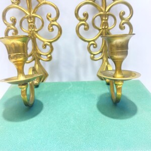 Puede incluir: Dos apliques de pared dorados y ornamentados. Cada aplique presenta un candelero con base circular y un dise&ntilde;o decorativo en espiral. Los apliques son de metal y est&aacute;n montados sobre una superficie verde claro.