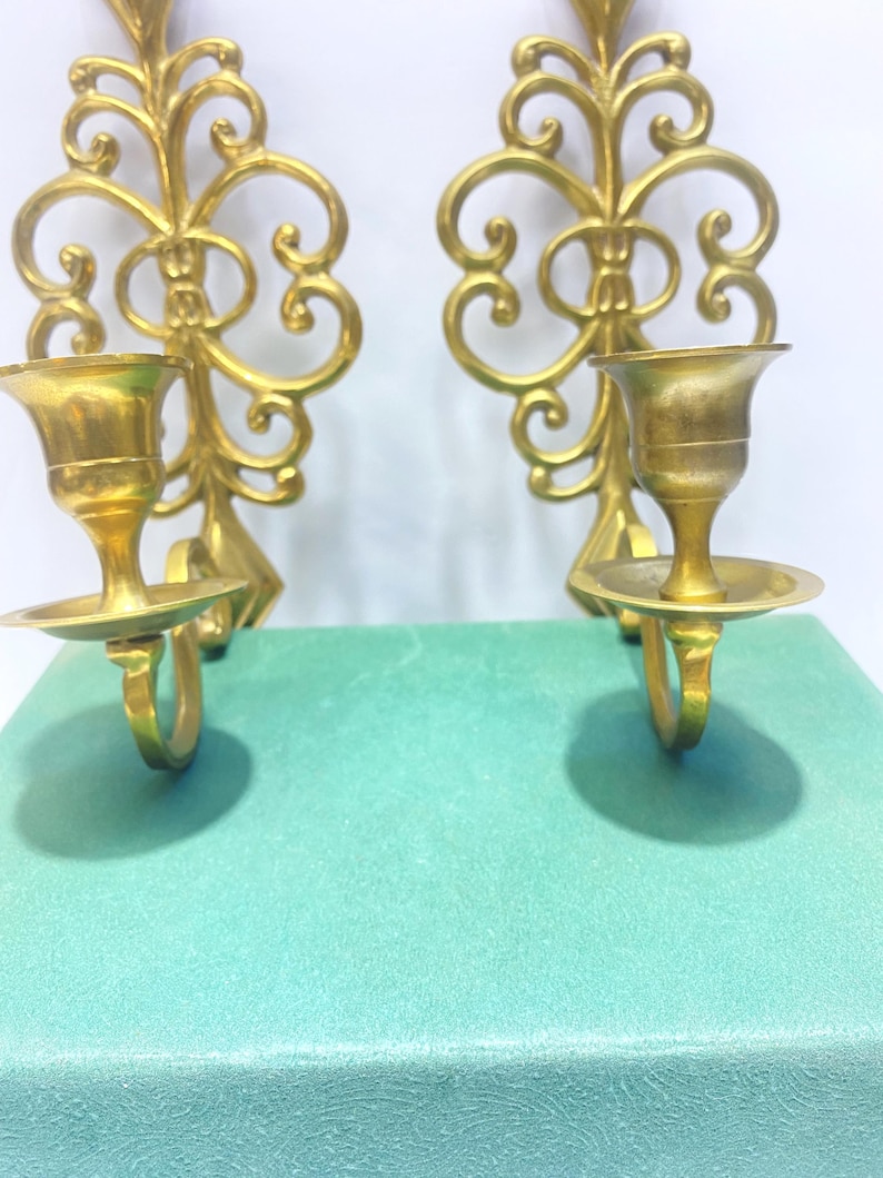 Puede incluir: Dos apliques de pared dorados y ornamentados. Cada aplique presenta un candelero con base circular y un dise&ntilde;o decorativo en espiral. Los apliques son de metal y est&aacute;n montados sobre una superficie verde claro.