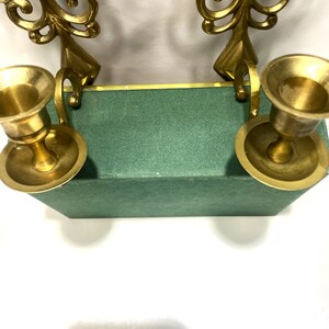 Puede incluir: Dos candelabros de lat&oacute;n con dise&ntilde;os ornamentados y arremolinados. Los vasos de las velas son redondos y se asientan sobre una base rectangular verde. Los candelabros son de color dorado c&aacute;lido y la base es verde apagado.