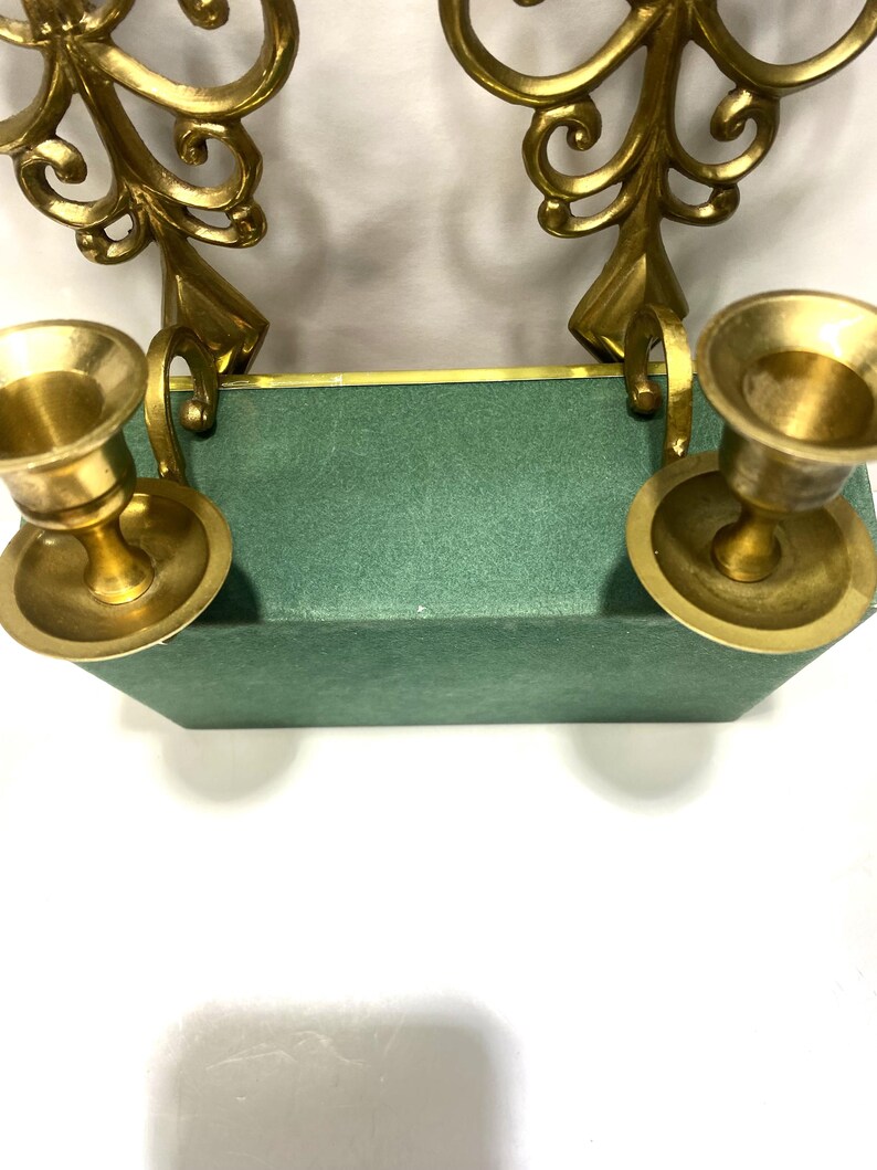 Puede incluir: Dos candelabros de lat&oacute;n con dise&ntilde;os ornamentados y arremolinados. Los vasos de las velas son redondos y se asientan sobre una base rectangular verde. Los candelabros son de color dorado c&aacute;lido y la base es verde apagado.