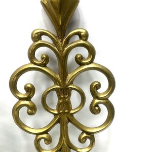 Puede incluir: Un elemento decorativo de metal dorado y ornamentado, con una parte superior en forma de diamante y un dise&ntilde;o sim&eacute;trico con volutas. La pieza tiene una est&eacute;tica vintage y es probablemente un componente decorativo arquitect&oacute;nico o de mobiliario.