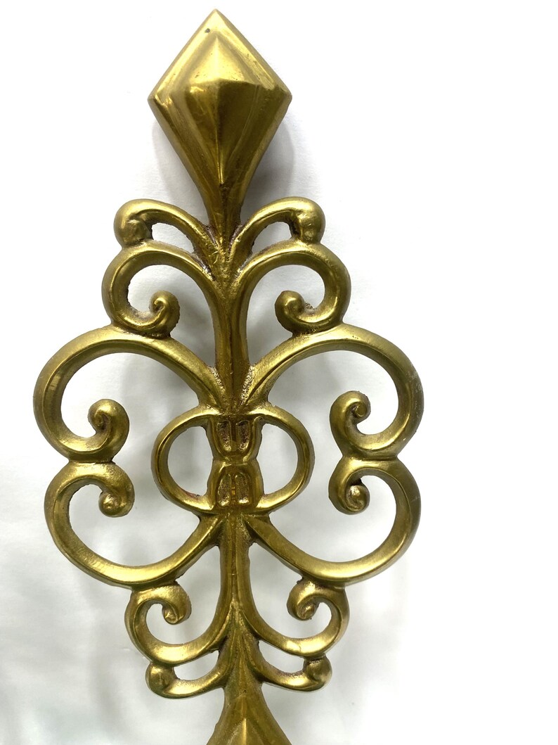 Puede incluir: Un elemento decorativo de metal dorado y ornamentado, con una parte superior en forma de diamante y un dise&ntilde;o sim&eacute;trico con volutas. La pieza tiene una est&eacute;tica vintage y es probablemente un componente decorativo arquitect&oacute;nico o de mobiliario.