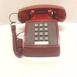 May include: A dark red landline telephone with a matching handset resting on the base. The device features a square keypad with black buttons, each marked with white numbers and letters, and a coiled cord connecting the handset to the base.
