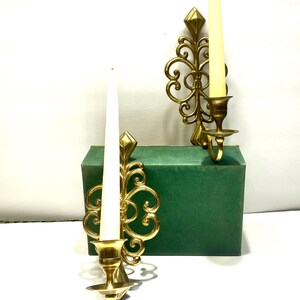 Puede incluir: Dos apliques de pared dorados y ornamentados con velas blancas. Cada aplique presenta un dise&ntilde;o decorativo en espiral y un acento en forma de diamante. Las velas son altas y delgadas. Los apliques se muestran sobre una caja verde.