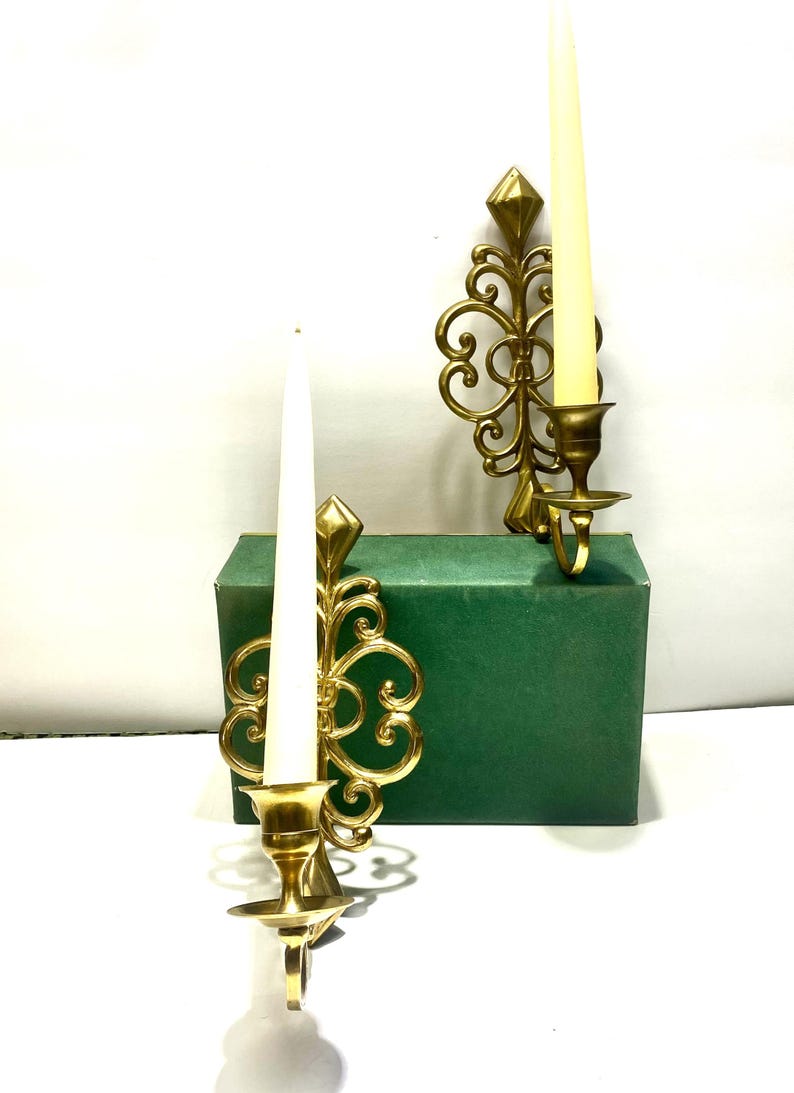 Puede incluir: Dos apliques de pared dorados y ornamentados con velas blancas. Cada aplique presenta un dise&ntilde;o decorativo en espiral y un acento en forma de diamante. Las velas son altas y delgadas. Los apliques se muestran sobre una caja verde.