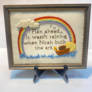 May include: A framed embroidery piece on off-white fabric featuring a colorful rainbow, a brown wooden ark, a yellow sun, and white clouds. The text reads: Plan ahead... it wasn't raining when Noah built the ark. The frame is dark brown wood.