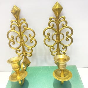 Puede incluir: Dos apliques de pared dorados y ornamentados. Cada aplique presenta una parte superior en forma de diamante, intrincados adornos y un portavelas con base circular. Los apliques se muestran sobre una superficie verde.