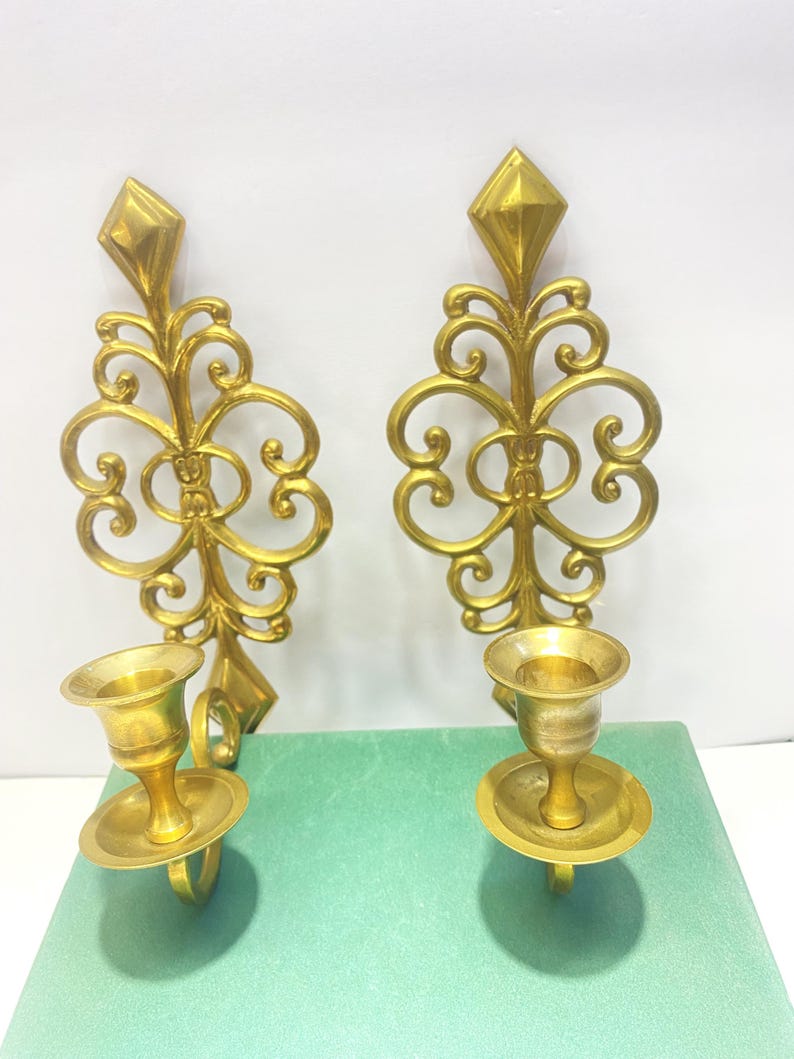 Puede incluir: Dos apliques de pared dorados y ornamentados. Cada aplique presenta una parte superior en forma de diamante, intrincados adornos y un portavelas con base circular. Los apliques se muestran sobre una superficie verde.