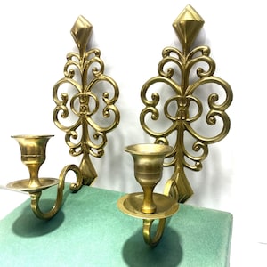 Puede incluir: Dos apliques de pared dorados y ornamentados. Cada aplique presenta un dise&ntilde;o decorativo en espiral con una parte superior en forma de diamante y un candelero. Los candeleros est&aacute;n unidos al soporte de pared con un brazo curvo.