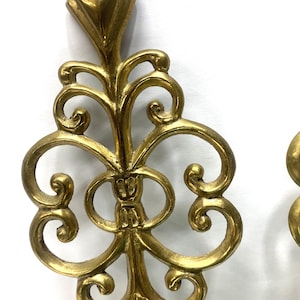 Puede incluir: Elemento decorativo de metal dorado y ornamentado, con una parte superior en forma de diamante y un dise&ntilde;o sim&eacute;trico con volutas. La pieza tiene una est&eacute;tica vintage y parece estar hecha de metal fundido. El dise&ntilde;o presenta l&iacute;neas curvas y formas circulares.
