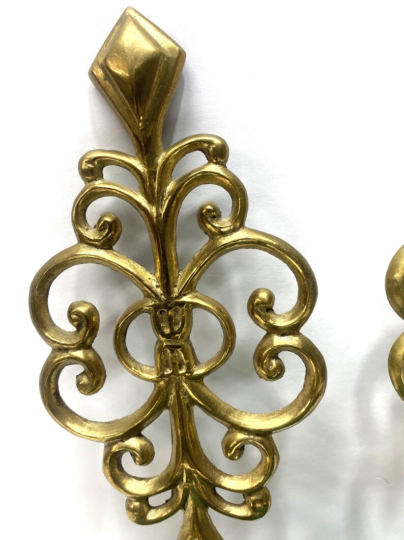 Puede incluir: Elemento decorativo de metal dorado y ornamentado, con una parte superior en forma de diamante y un dise&ntilde;o sim&eacute;trico con volutas. La pieza tiene una est&eacute;tica vintage y parece estar hecha de metal fundido. El dise&ntilde;o presenta l&iacute;neas curvas y formas circulares.