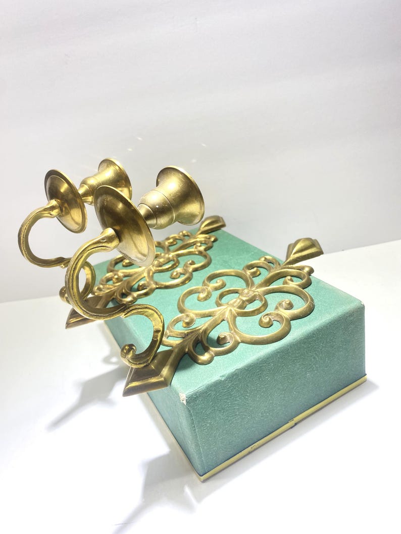 Puede incluir: Dos candelabros de lat&oacute;n dorado adornados con un dise&ntilde;o floral decorativo. Los candelabros son de color dorado y presentan brazos curvos y copas en forma de campana. Los candelabros est&aacute;n sobre una caja de color turquesa.