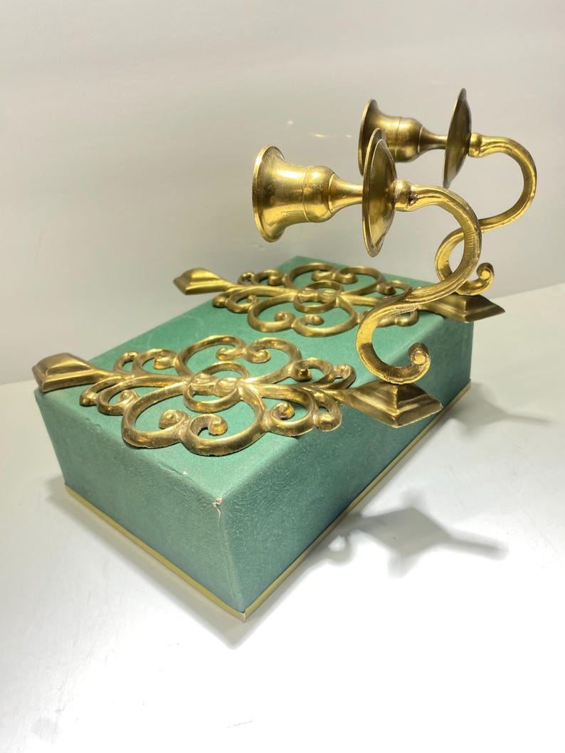 Puede incluir: Un par de candelabros de lat&oacute;n dorado con adornos de volutas y brazos curvos. Montados sobre una caja rectangular de color verde azulado con ribetes dorados. El dise&ntilde;o es cl&aacute;sico y elegante, adecuado para la decoraci&oacute;n del hogar.