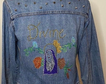 May include: A light blue denim jacket with silver studs on the shoulders and a colorful embroidered design on the back. The design features a woman's face with the word "Divine" above it.