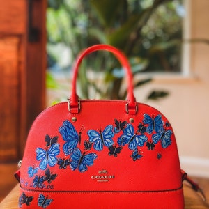 May include: A red leather satchel handbag with a gold Coach logo and a design of blue butterflies. The bag has a top handle and a detachable strap. The text "Take a chance" is written on the bag.