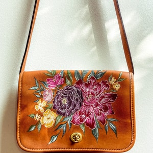 May include: A brown leather shoulder bag with a floral design painted on the front. The flowers are in shades of pink, purple, and yellow.