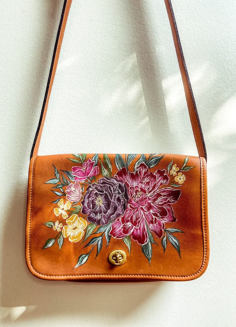 May include: A brown leather shoulder bag with a floral design painted on the front. The flowers are in shades of pink, purple, and yellow.