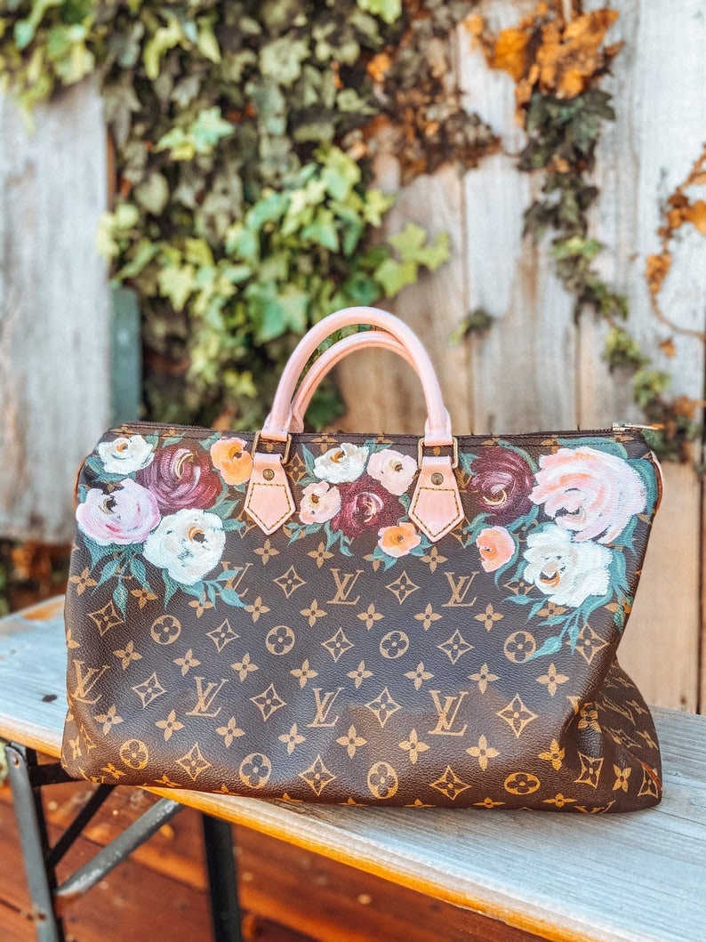 May include: A brown and tan handbag with a floral pattern painted on the front. The bag has a pink leather handle and gold hardware.