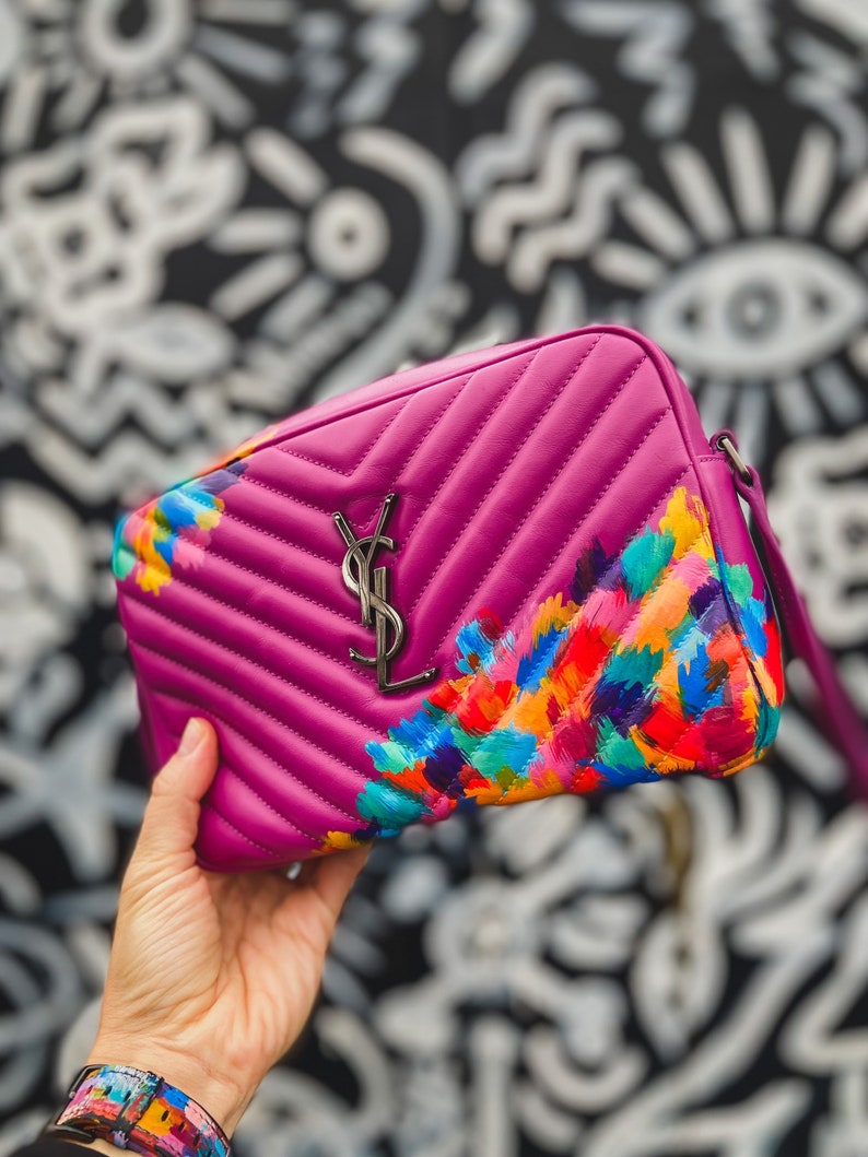 May include: A pink quilted leather handbag with a silver YSL logo and colorful abstract floral design.