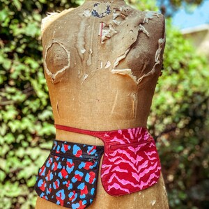 May include: A brown leather belt with two attached pouches. The pouch on the left has a black, blue, and red leopard print. The pouch on the right has a pink and red zebra print.