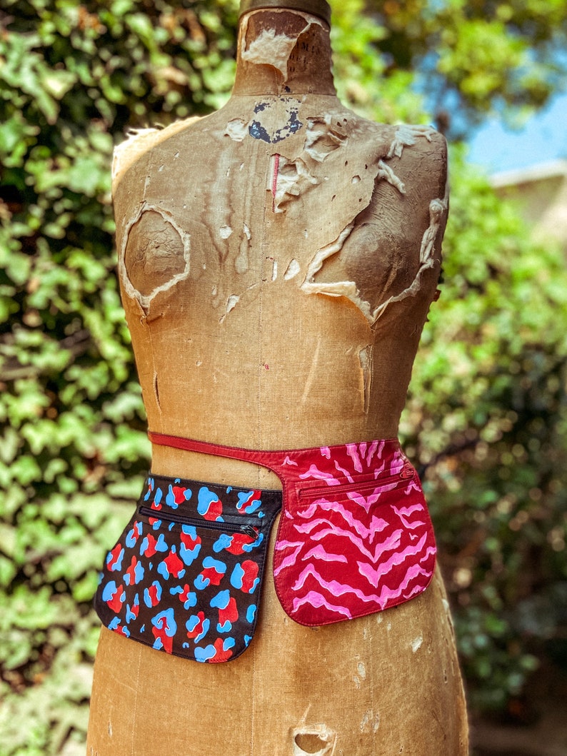 May include: A brown leather belt with two attached pouches. The pouch on the left has a black, blue, and red leopard print. The pouch on the right has a pink and red zebra print.