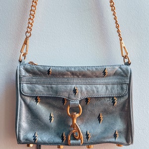 May include: A gray leather crossbody bag with a gold chain strap and a gold clasp. The bag has a black lightning bolt pattern.