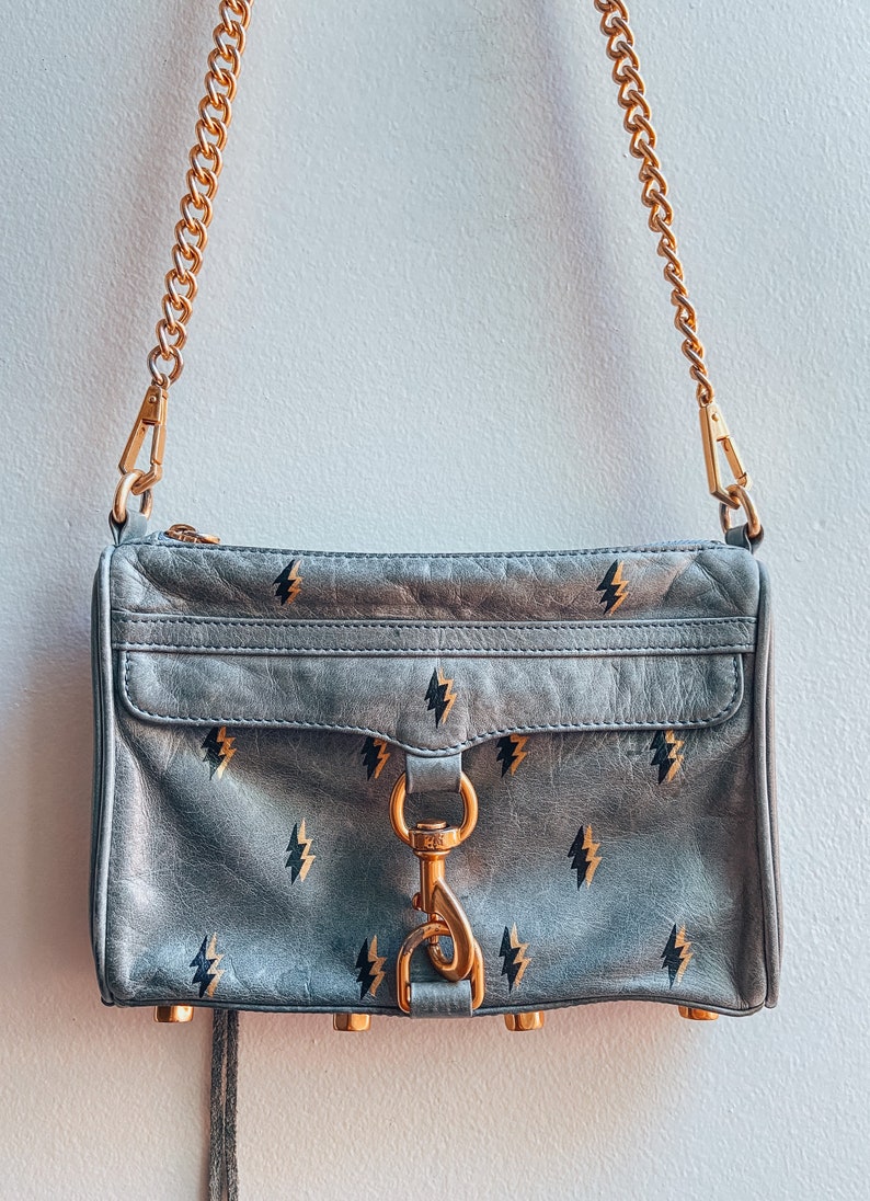 May include: A gray leather crossbody bag with a gold chain strap and a gold clasp. The bag has a black lightning bolt pattern.