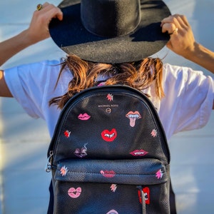May include: Black leather backpack with a pink and red lips and teeth design. The backpack has a zipper closure and a small pocket on the front. The brand name "MICHAEL KORS" is printed on the front of the backpack.