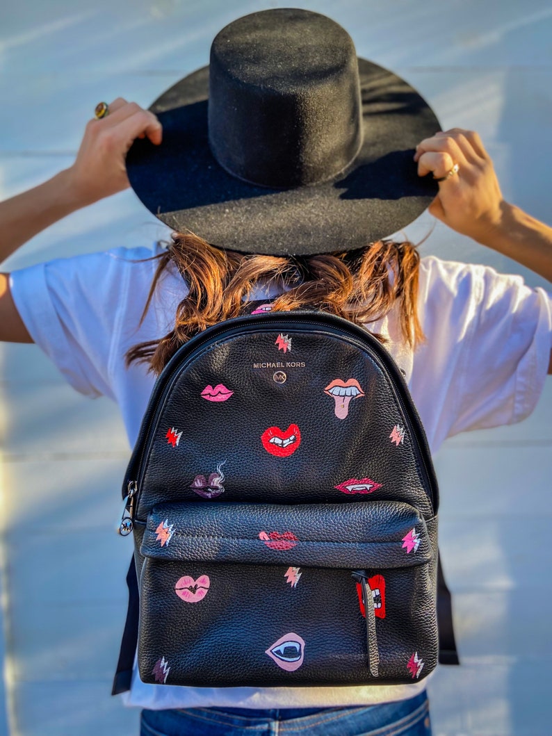 May include: Black leather backpack with a pink and red lips and teeth design. The backpack has a zipper closure and a small pocket on the front. The brand name "MICHAEL KORS" is printed on the front of the backpack.