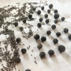 May include: An overhead shot displays a collection of small, dark gray beaded spheres and loose beads scattered on a white, textured surface. The spheres vary in size, creating a textured, organic pattern. The beads are metallic.
