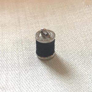 May include: A silver charm in the shape of a spool of thread. The spool is black and has a silver loop at the top.