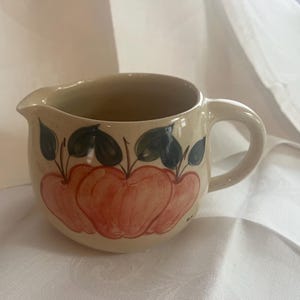 May include: A beige ceramic pitcher with a handle, featuring a hand-painted design of red apples and green leaves.