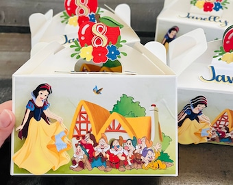 May include: White and yellow favor boxes with a Snow White and the Seven Dwarfs theme. The boxes have a red apple with the number 8 on top. The boxes are decorated with a picture of Snow White and the Seven Dwarfs in front of their cottage.