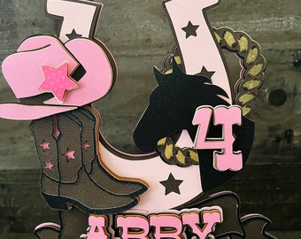 May include: Pink and brown cake topper with a horseshoe, a cowboy hat, a boot, a horse head, a star, and the name Abby. The number 4 is also included on the topper.