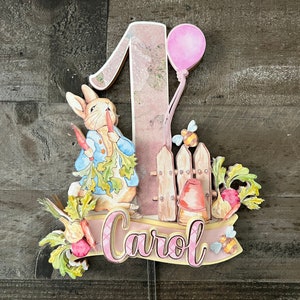 May include: A pink and white wooden cake topper featuring a rabbit holding carrots, a fence, a pink balloon, and the name "Carol".