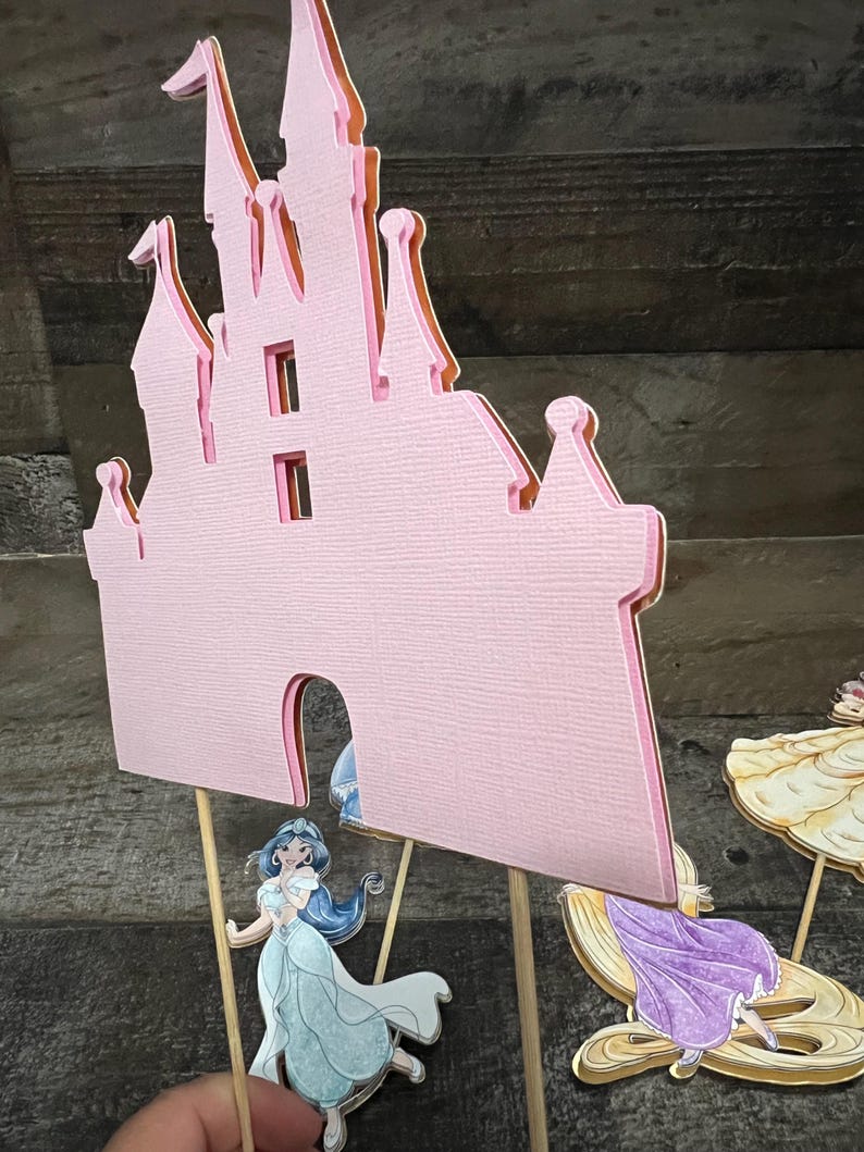 May include: A pink paper castle cutout with a light blue princess figure in front of it. The princess has long dark hair and is wearing a light blue dress. There are two other princess figures in the background.