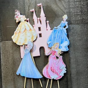 May include: Four Disney princess cake toppers featuring Belle, Cinderella, Elsa, and Aurora. Each princess is depicted in her signature gown and is attached to a wooden stick. The princesses are arranged in front of a pink castle cutout.
