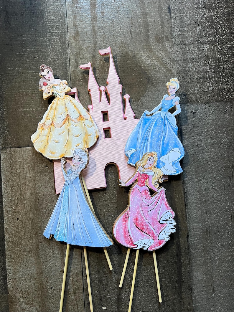 May include: Four Disney princess cake toppers featuring Belle, Cinderella, Elsa, and Aurora. Each princess is depicted in her signature gown and is attached to a wooden stick. The princesses are arranged in front of a pink castle cutout.