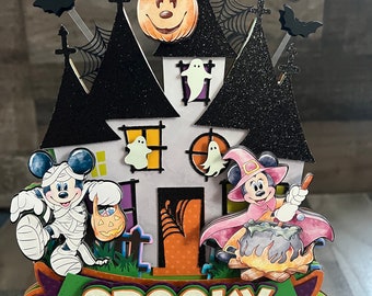 May include: A Halloween-themed decoration featuring a house with black glittered roofs, bats, and a pumpkin-shaped Mickey Mouse. Mickey Mouse and Minnie Mouse are dressed in Halloween costumes. The word "SPOOKY" is displayed on a banner.