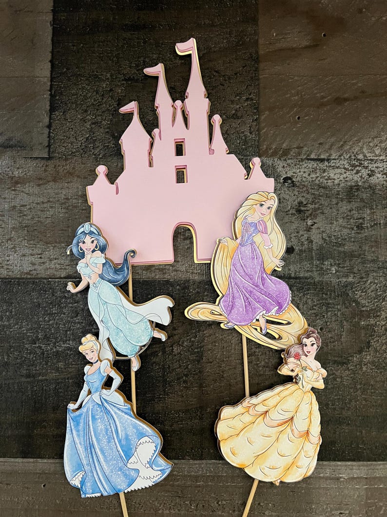 May include: A set of four princess character cutouts, including Cinderella, Belle, Rapunzel, and Jasmine, with a pink castle cutout. The cutouts are on wooden sticks and are perfect for a princess-themed party.