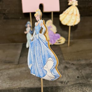 May include: A blue and white Cinderella cake topper with a sparkly blue dress and a golden trim. The topper is on a wooden stick.